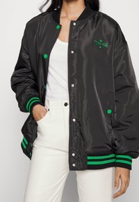 Svart satin bomberjacka med gröna randiga ribbade manschetter och nederkant. Broderad logotyp på vänster bröst, tryckknappsstängning.