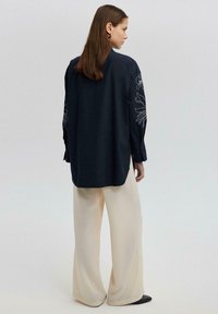 Camicia a maniche lunghe di colore navy con ricami floreali sulle maniche, abbinata a pantaloni a gamba larga color crema e scarpe nere. Texture del tessuto liscia.