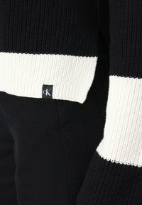 Maglione a righe bianco e nero in maglia con texture a coste, dotato di un orlo bianco e di un piccolo etichetta del marchio nell'angolo inferiore.