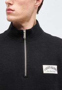 Άνδρας που φοράει μαύρο φούτερ Calvin Klein με φερμουάρ, με ψηλό γιακά, το φερμουάρ είναι μερικώς κλειστό κοντά στον λαιμό.