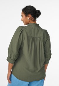 Olivgrön, lätt blus med puffärmar, rynkad halsringning och veck baktill. Har en slät textur och en avslappnad passform.