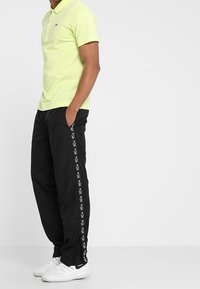 Neon žluté polo tričko, černé kalhoty s bílým logem na lemu a bílé tenisky. Polo má límec; kalhoty mají volný střih.