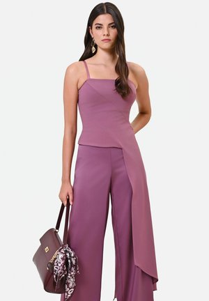 Femme portant un top asymétrique mauve sans manches et un pantalon large assorti, tenant un sac à main violet foncé avec un foulard à motif en accessoire.