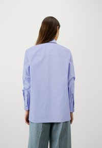 Camicia viola chiaro a maniche lunghe con colletto strutturato, polsini abbottonati e una texture di cotone morbido, abbinata a pantaloni in denim a gamba larga.