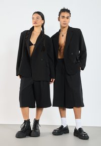 Black oversized blazers, relaxed fit shorts, chunky black boots with laces, and loafers. Minimalist design with emphasis on bold shapes.