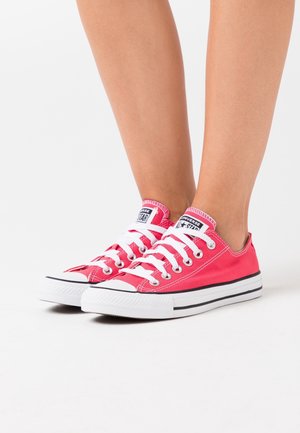 Zapatillas de lona rojas con cordones blancos y suelas de goma. Presentan una puntera redondeada y una franja negra a lo largo de la media suela. Marca icónica en la lengüeta.