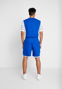 Hombre de pie dando la espalda, vistiendo una camisa de manga corta azul y blanca, pantalones cortos azules y zapatillas deportivas blancas, sobre un fondo liso.