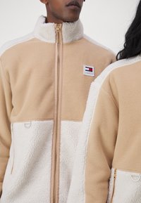 Giacca in pile con un design a blocchi di colore beige e bianco. Collo alto, chiusura con zip e patch del logo sul petto. Morbida al tatto.