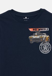 Tumedate puuvillane T-särk, millel on graafika võidusõiduautost, mille ülaosas on tekst "HOT WHEELS" ja allosas ringikujuline logo.