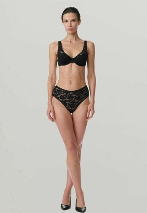 Kvinna som bär svart spetsbh och högmidjade trosor, står med korsade ben framför en enhetlig ljusgrå bakgrund.