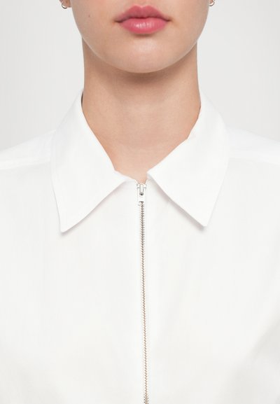 Witte ritsblouse met een klassieke kraag, een gladde textuur en zilveren ritsdetails aan de voorkant.