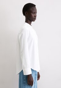 Biała koszula z długim rękawem i stójką, ozdobiona pionowymi paskami oraz zapięciem na guziki z przodu. Połączona z jasnoniebieskimi dżinsami.