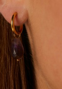 Boucle d'oreille en or avec un pendentif en améthyste facettée de couleur violette. La pierre a la forme d'une goutte et met en valeur des teintes violettes claires et une texture naturelle.
