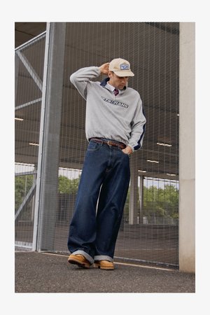 Grå hettegenser med marineblå detaljer og logo, blå vide jeans, brune sko, og en beige caps. Bakgrunnen har en metallnettkonstruksjon.