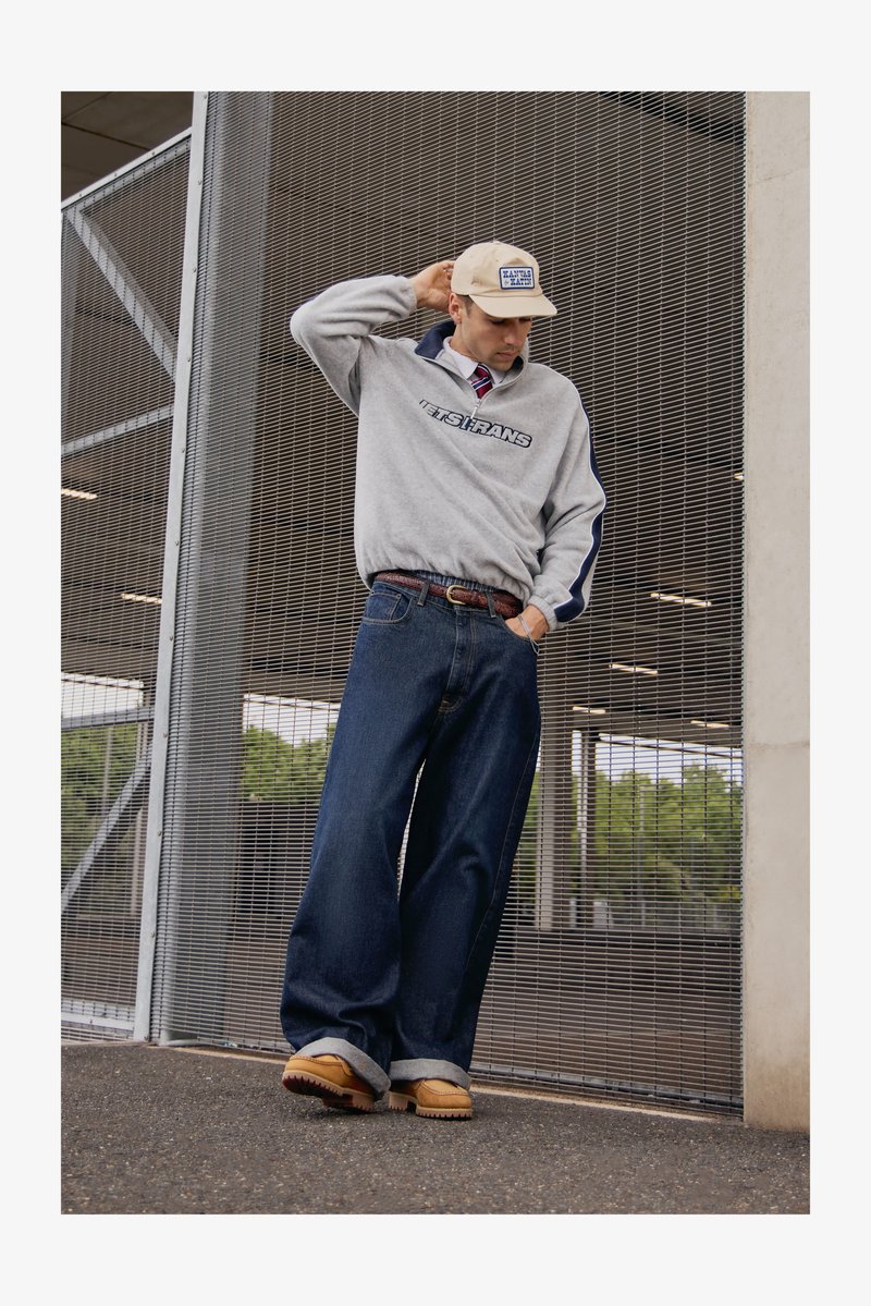 Grå hettegenser med marineblå detaljer og logo, blå vide jeans, brune sko, og en beige caps. Bakgrunnen har en metallnettkonstruksjon.