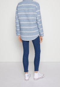 Blårandig flanellskjorta med krage, i kombination med åtsittande blå jeans och vita hög-toppsneakers. Har uppvikta ärmar och rundad fåll.