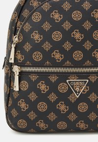 Zaino nero progettato con un motivo di logo beige ripetuto, dotato di hardware per cerniere in oro metallico e un accento con logo triangolare del marchio.