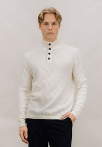 Hvit strikket genser med høy ribbestrikket krage, med et vevd mønster og fem mørke knapper ved halsen. Kombinert med mørke bukser.