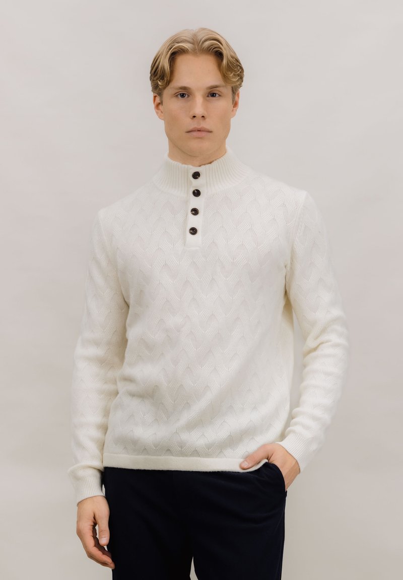 Hvit strikket genser med høy ribbestrikket krage, med et vevd mønster og fem mørke knapper ved halsen. Kombinert med mørke bukser.