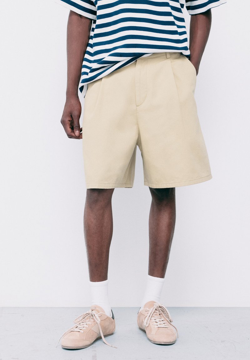 Homme portant un short beige mi-genou, des chaussettes blanches, des baskets beiges et une chemise à manches courtes rayée bleu et blanc, debout contre un mur blanc.