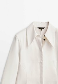 Chemise blanc cassé à manches longues avec col large et pointu et patte de boutonnage cachée, deux boutons noirs visibles près du col.