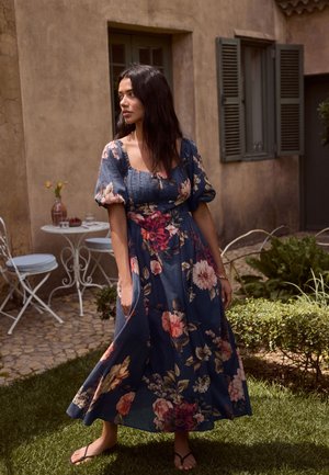 Nő kék virágos ruhában, mezítláb áll a fűben, közel egy kőerkélyhez, fehér fém asztallal és székekkel, bézs fal mellett.