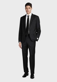 Completo nero su misura con rever a punta, camicia bianca e cravatta nera. Scarpe nere con finitura lucida, dotate di un dettaglio di fazzoletto da taschino.