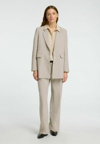Traje ajustado de color beige claro que incluye una chaqueta larga y estructurada con bolsillos de parche, combinada con pantalones acampanados a juego y una camisa de botones beige.
