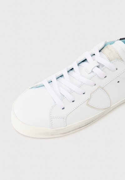 Baskets blanches avec tige en cuir lisse, bout rond, lacets blancs et détails bleu clair sur la languette. Design minimaliste avec accents latéraux.