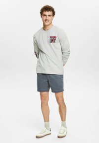 Lys grå langærmet skjorte med "NY" grafik på brystet, parret med mørkegrå shorts og hvide sneakers med brune accenter.