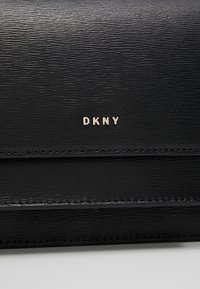 Černý kožený povrch s texturou a malými zlatými písmeny tvořícími nápis "DKNY" uprostřed blízko prošitého švu.