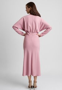 Różowa sukienka z długim rękawem, z wcięciem w talii i zwiewnym dołem. Gładki, miękki materiał oraz prosty krój. Widok z tyłu ukazuje pełną długość.
