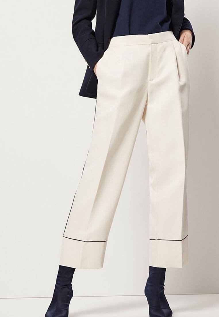 Personne portant un pantalon crème à jambes larges avec bordure noire, un haut bleu marine et des bottines bleu marine à talons, debout les mains dans les poches.