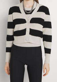 Cárdigan de punto a rayas negras y beige con escote en V, tres botones y puños acanalados. Diseño corto, combinado con pantalones ajustados negros.