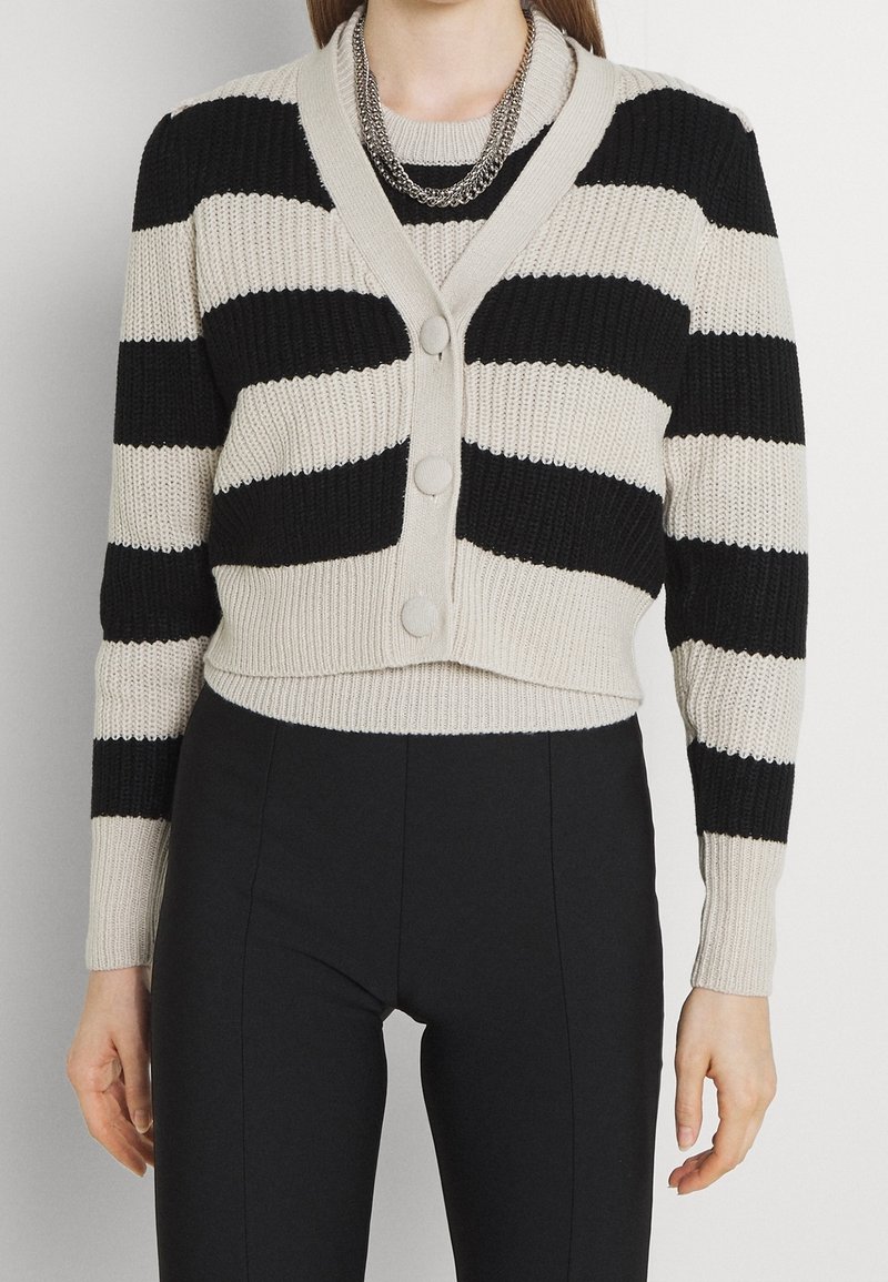 Cárdigan de punto a rayas negras y beige con escote en V, tres botones y puños acanalados. Diseño corto, combinado con pantalones ajustados negros.