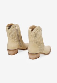 Botines marrones con perforaciones en los laterales, textura suave, cierre de cremallera lateral y un tacón bajo apilado. Suela plana y diseño de puntera redondeada.