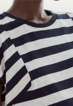 Nahaufnahme eines Oberkörpers, der ein kurzärmeliges marineblau-weiß gestreiftes Shirt mit rundem Ausschnitt trägt.