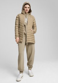 Lys beige pufferjakke med hætte, lynlås lukning og horisontal quiltet tekstur, båret over et matchende beige sæt og hvide sneakers.