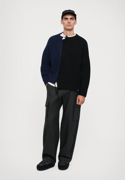 Chunky knit sweater with a blue textured half and a solid black half, paired with loose black cargo pants and black embroidered slip-ons.