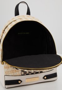 Sac à dos rond avec un extérieur tissé beige et noir, des fermetures éclair dorées et un intérieur en tissu noir avec une étiquette de marque à l'intérieur.