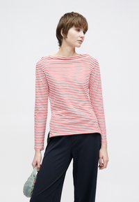 Czerwona i biała pasiasta bluzka z długim rękawem i dekoltem w łódkę, zestawiona z ciemnogranatowymi spodniami, trzymana jedną ręką. Gładka tekstura, lekki materiał.