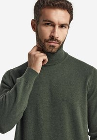 Mörkgrön rollkragetröja, stickad textur, figurnära design, mjukt tyg, ribbad krage och ärmslut, bärs med hand på nacken, neutral bakgrund.