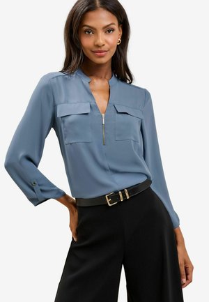 Blusa azzurra chiara con colletto, due tasche sul petto e dettaglio con zip. Maniche lunghe, tessuto liscio, abbinata a una cintura nera e pantaloni.
