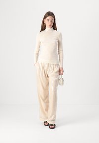 Lichtbeige doorschijnende turtleneck-top gecombineerd met losse, met pailletten bezette crèmekleurige broeken. De outfit heeft een relaxte pasvorm en zwarte sandaaltjes met banden.