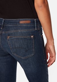 Los jeans de mezclilla azul oscuro presentan un parche de cuero en la cintura, acentuado con costuras en contraste y un diseño en el bolsillo trasero.