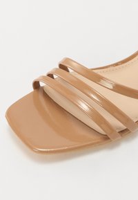 Ruskea neliön muotoinen sandaali, jossa on kiiltävä hihnadesign ja sileä beige sisustus. Yläosa sisältää neljä kiiltävää, ristikkäin kulkevaa hihnaa.