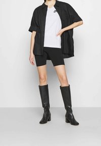 Person som bär svarta knälånga stövlar, svarta cykelshorts, vit Nike-t-shirt och en lös svart skjorta med knappar, står mot en enfärgad bakgrund.