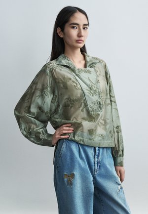 Przezroczysta oliwkowozielona bluzka z kwiatowym haftem, luźnego kroju, z kołnierzykiem. W zestawieniu z jasnoniebieskimi dżinsami z motywami zwierzęcymi.