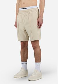 Beige atletiske shorts med en taljebånd med logo og subtile folder, lavet af glat stof, båret med hvide sokker med logo og beige sneakers.