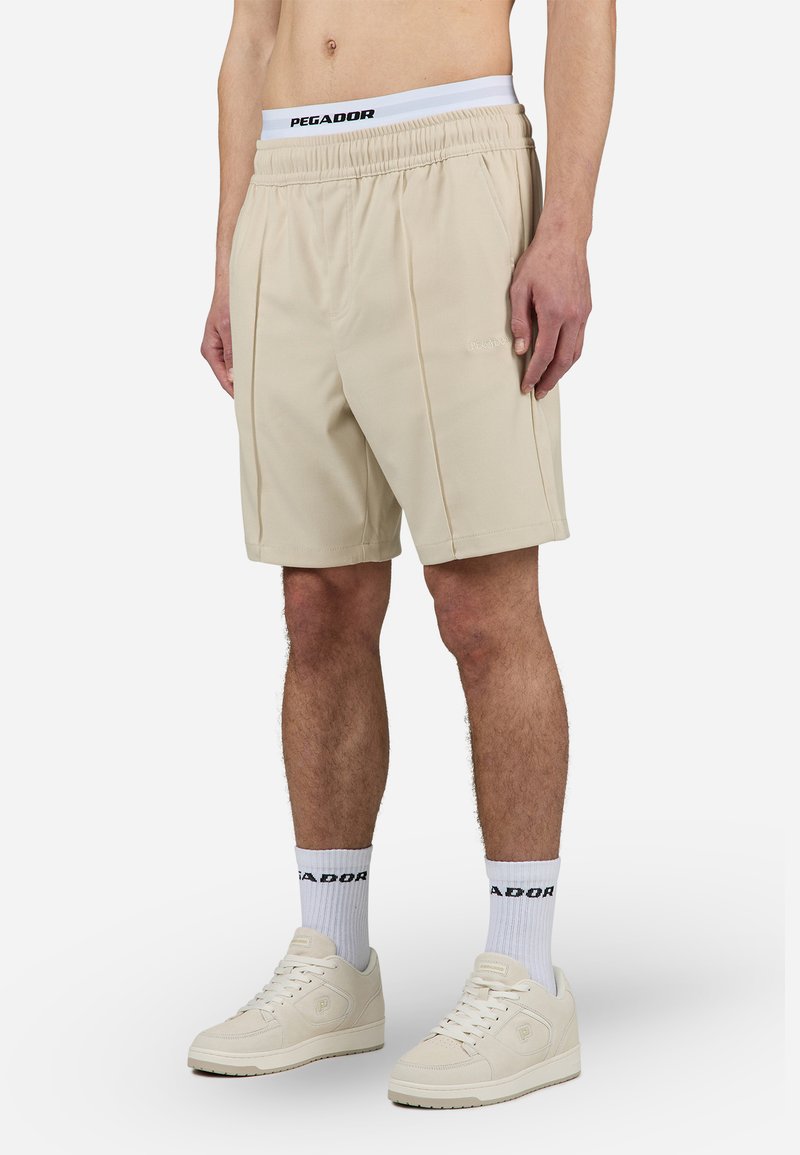 Beige atletiske shorts med en taljebånd med logo og subtile folder, lavet af glat stof, båret med hvide sokker med logo og beige sneakers.